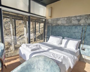 A bedroom with a stone wall, a bed with white linens, and a view of a rocky landscape through large windows. - Stunning Cliff-edge 2-bedroom Chalet in Karimabad (Karimabad)