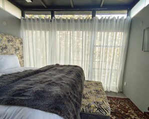 Ein Zimmer mit einem Bett, einer gemusterten Bank, durchscheinenden Vorhängen und Blick auf Bäume durch das Fenster.