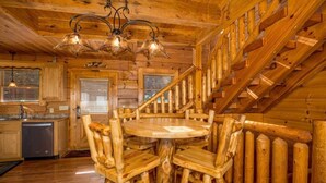 A rustic wooden interior with a round wooden table surrounded by chairs, a staircase, and a kitchen area.