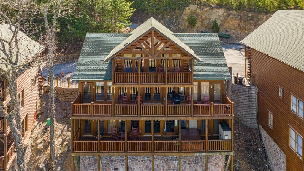 Une cabane en rondins à plusieurs étages, dotée d’un balcon à chaque niveau, entourée d’arbres et située au pied d’une colline rocheuse.