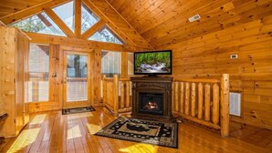 A log cabin interior with a fireplace, a television, and large windows.