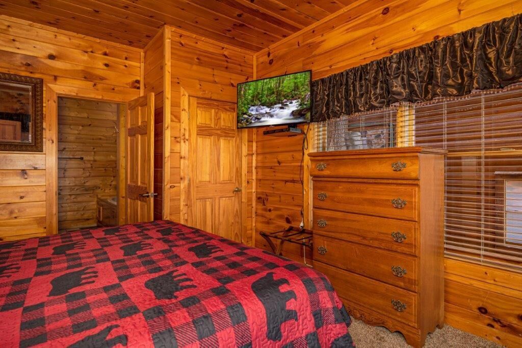 A wooden cabin bedroom with a bed, a dresser, a television, and a window with blinds.