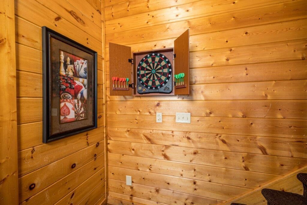 A wooden wall with a dartboard and darts, a framed picture of playing cards, and a small cabinet.