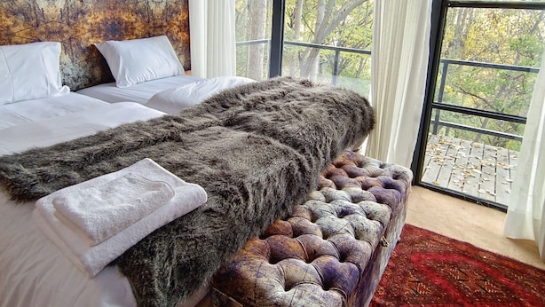 A bedroom with a large bed, a fur throw, a tufted bench, and a view of trees through large windows. - Kalp With Forest View- Cloud Nine and a Half Hunza (Karimabad)