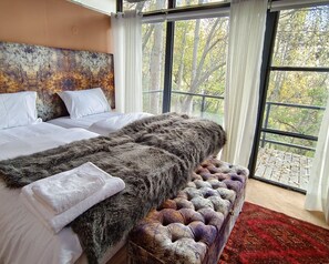 A bedroom with a large bed, a fur throw, a tufted bench, and a view of trees through large windows. - Kalp With Forest View- Cloud Nine and a Half Hunza (Karimabad)