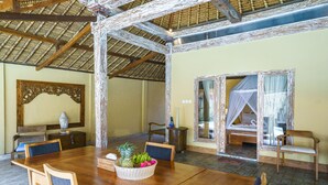 A wooden dining table with chairs, a fruit basket, and a bed with a mosquito net in a room with a tiled floor and a thatched roof. - Villa Matahari (Gili Meno)