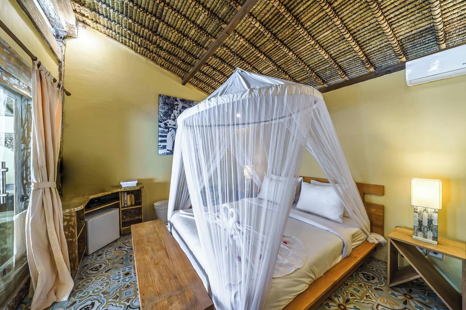 A room with a bed under a mosquito net, a bedside table with a lamp, a small shelf with books, and a patterned floor.