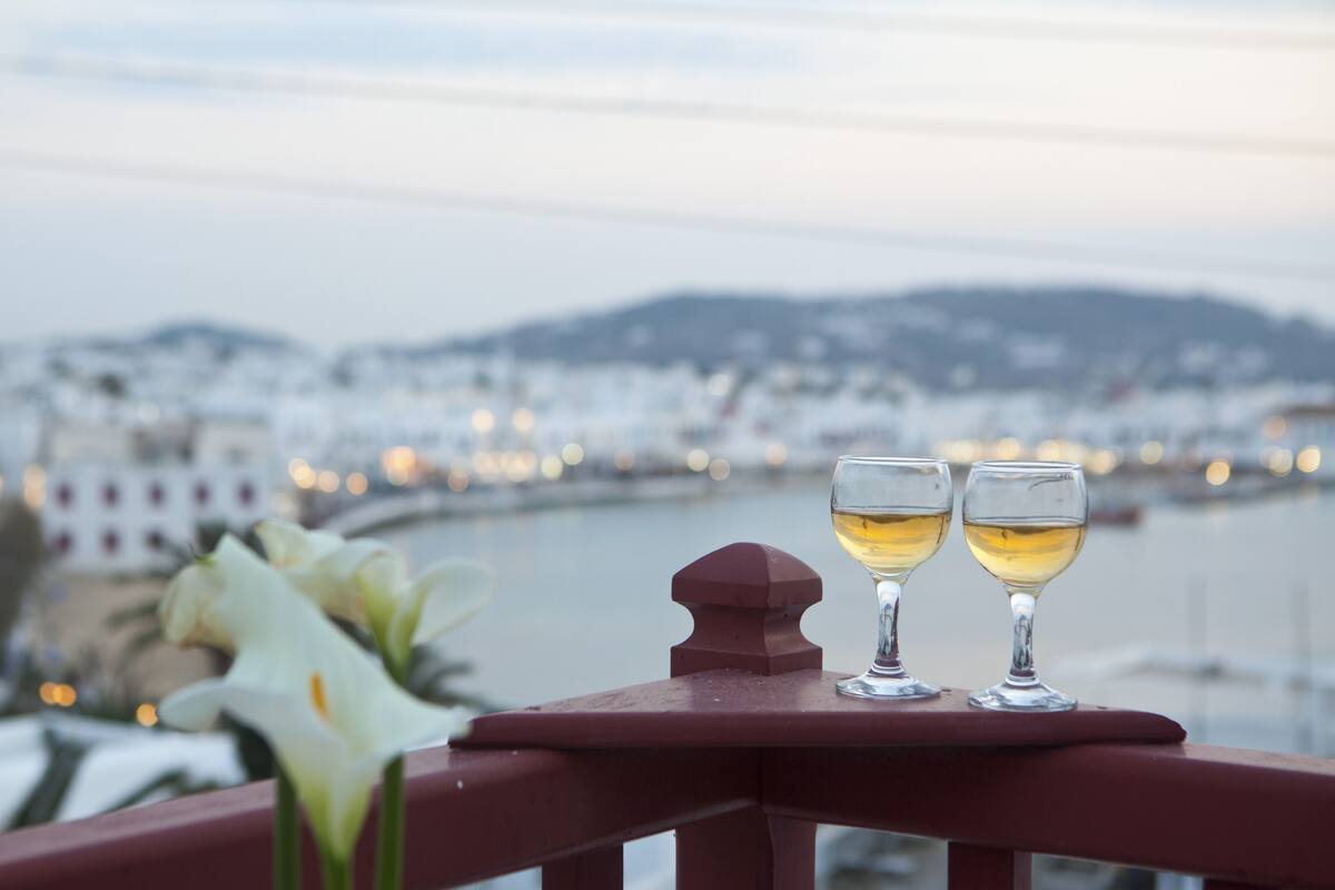 Dos copas de vino blanco sobre una barandilla de balcón con vistas a un paisaje urbano y una masa de agua.