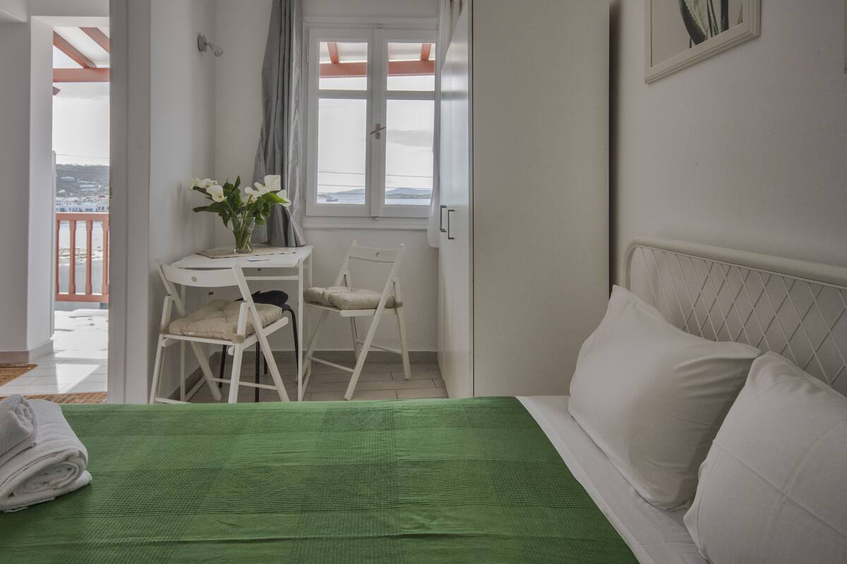 Un dormitorio con colcha verde, almohadas blancas, un escritorio pequeño con silla y una ventana con cortinas.