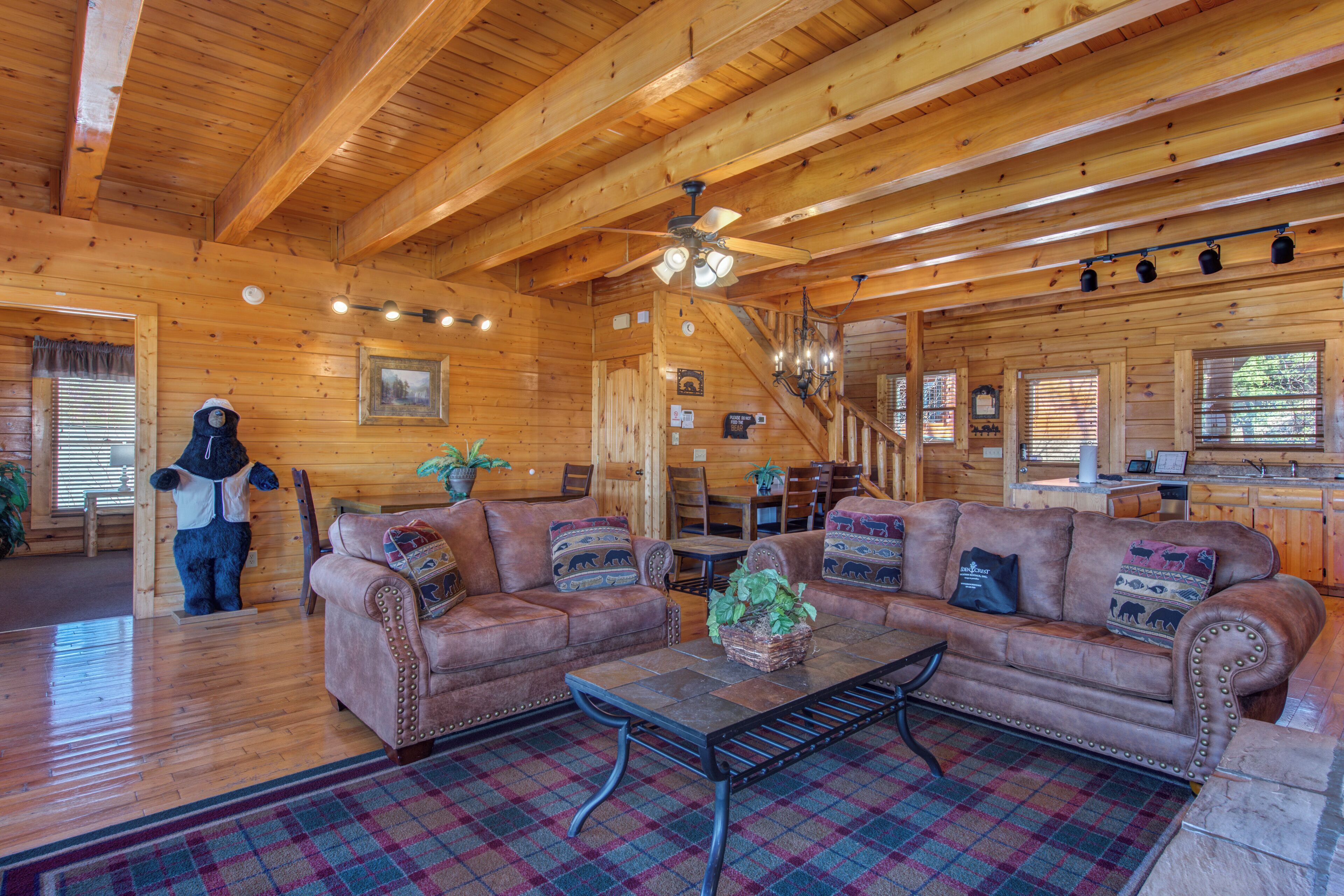 A cozy log cabin interior with a wooden ceiling, a large brown leather sofa, a plaid rug, and a dining area with wooden tables and chairs.