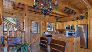 A wooden cabin interior with a dining table, chairs, a chandelier, a kitchen with appliances, and a staircase. - Royal Vista Lodge (Sevierville)