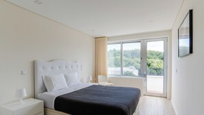Un dormitorio moderno con una cama grande, una mesita de noche y un escritorio junto a la cama, y una ventana con vistas a zonas verdes.