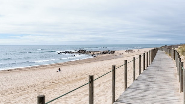 Una pasarela de madera bordea una playa arenosa con un promontorio rocoso en el agua.