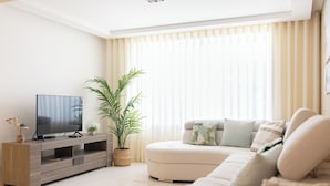 Una sala de estar moderna con un televisor de pantalla plana, un sofá gris, una planta grande en maceta y una ventana con persianas verticales.
