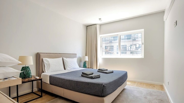 A neatly arranged bedroom with a bed, bedside tables, a window with curtains, and a view of the cityscape. - Lu | Localização Central, Vista Rio and Nespresso (Vila Nova de Gaia)