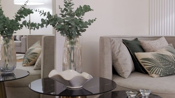 Een moderne woonkamer met een bank, twee salontafels met glazen bladen en gouden poten, en decoratieve vazen met groene planten.