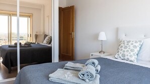 A bedroom with a large bed, a nightstand with a lamp, a wooden door, and a view through a sliding door. - Porto Gaia River View (Vila Nova de Gaia)