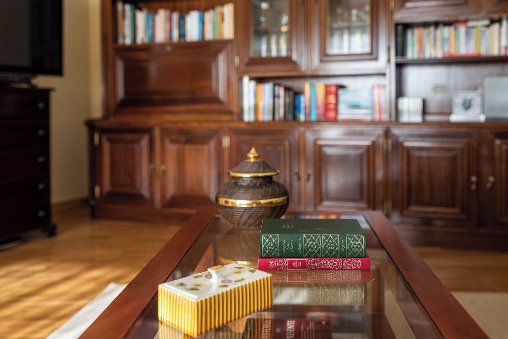 A wooden bookshelf filled with books, a decorative ceramic jar, and a set of books on a glass table.