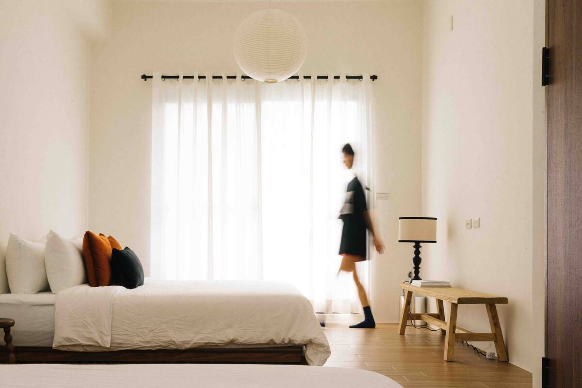 Quarto com cama, almofadas, mesa de cabeceira, abajur e janela com cortinas.