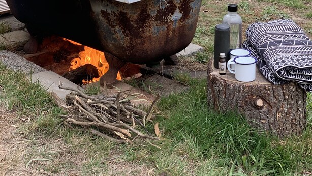 Un décor extérieur rustique avec une grande baignoire rouillée remplie d’eau, entourée d’un foyer, et divers articles de camping, comme une bouteille d’eau, une tasse et une couverture, disposés sur le tronc d’un arbre.