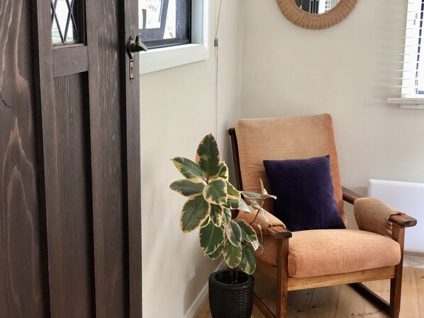A room with a wooden door, a potted plant, a wooden chair with a cushion, and a round mirror on the wall.