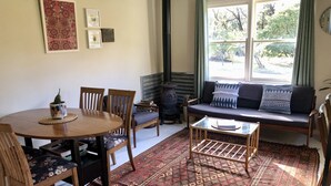 Un salon chaleureux avec une table ronde en bois, deux chaises en bois, un tapis à motifs, un canapé avec des coussins et une grande fenêtre avec des rideaux.