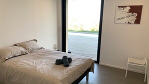 Een slaapkamer met een bed, kussens, een nachtkastje, een lamp en een witte stoel. Een deur leidt naar een buitenruimte met uitzicht op groen.