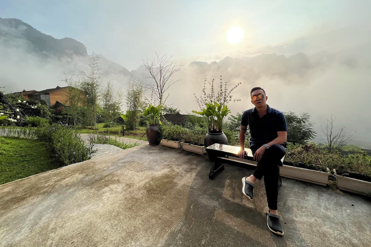 A man sitting on a concrete platform with potted plants, overlooking a mountainous landscape with a clear sky.