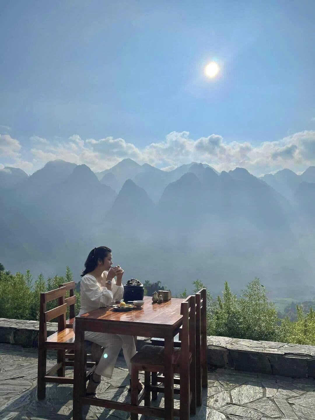 A person sitting at a wooden table with a view of mountains and a bright sun in the background.