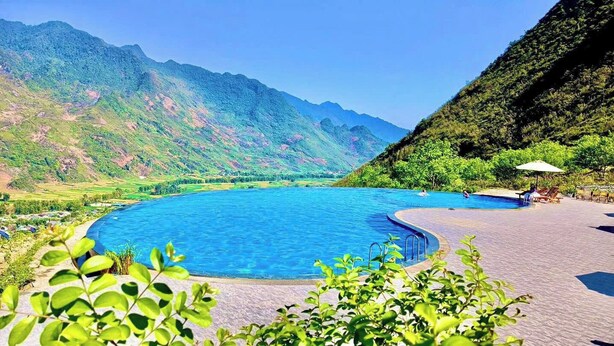 A swimming pool with a mountain backdrop, surrounded by lush greenery and a paved walkway. - H'mong Village Resort Ha Giang (Lung Tam)