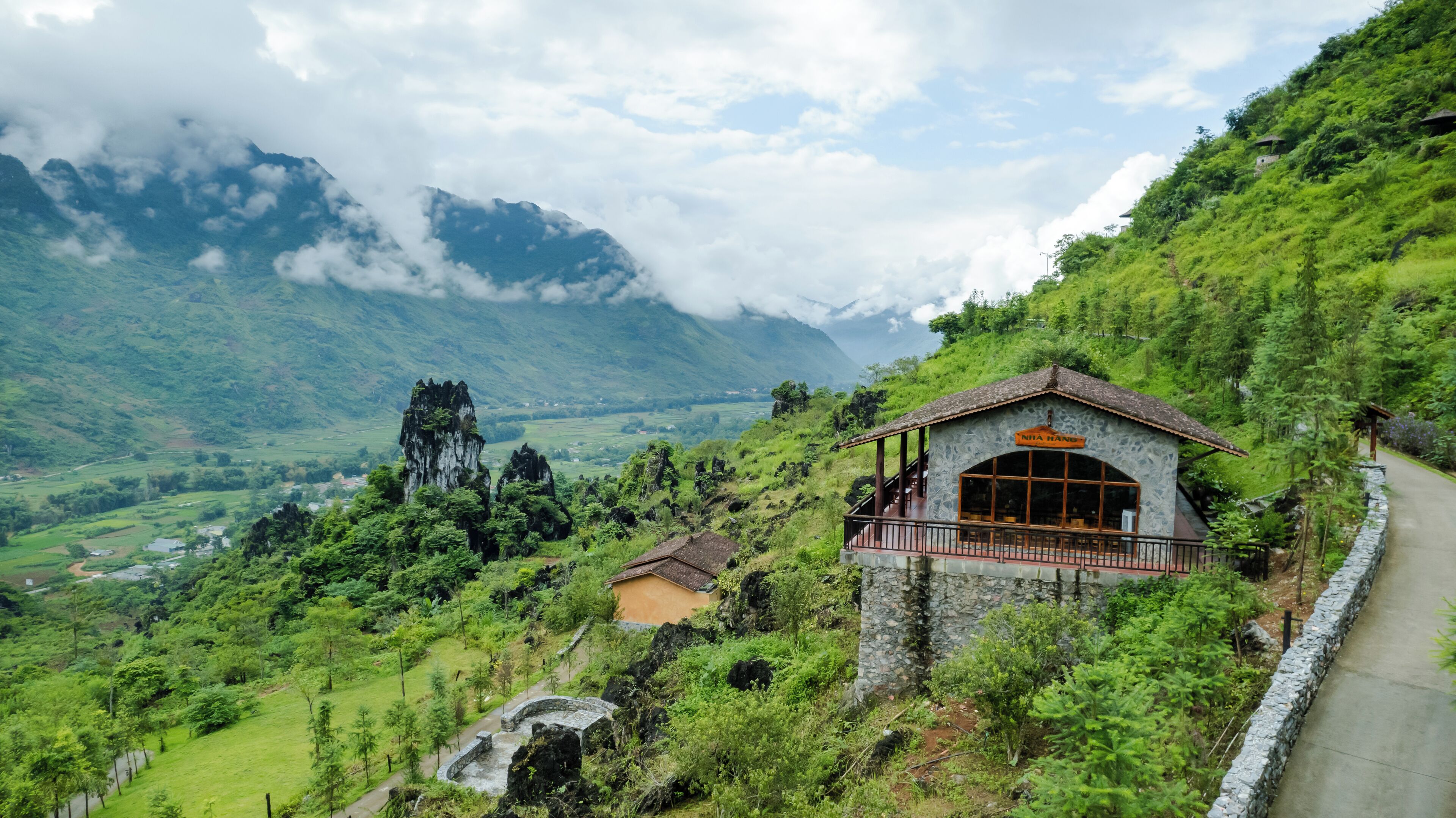 A stone building with a balcony, surrounded by lush greenery and mountains in the background.