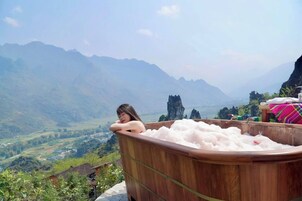 Uma pessoa relaxando em uma banheira de madeira com vista panorâmica para montanhas e um vale.