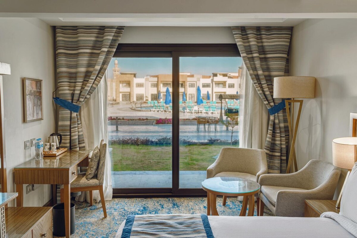Quarto de hotel com cama, duas poltronas, uma mesinha, escrivaninha e vista para a piscina e edifícios.