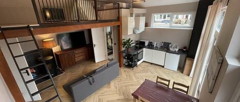 A loft-style apartment with a dining area, kitchen, and a loft bedroom with a TV and storage.