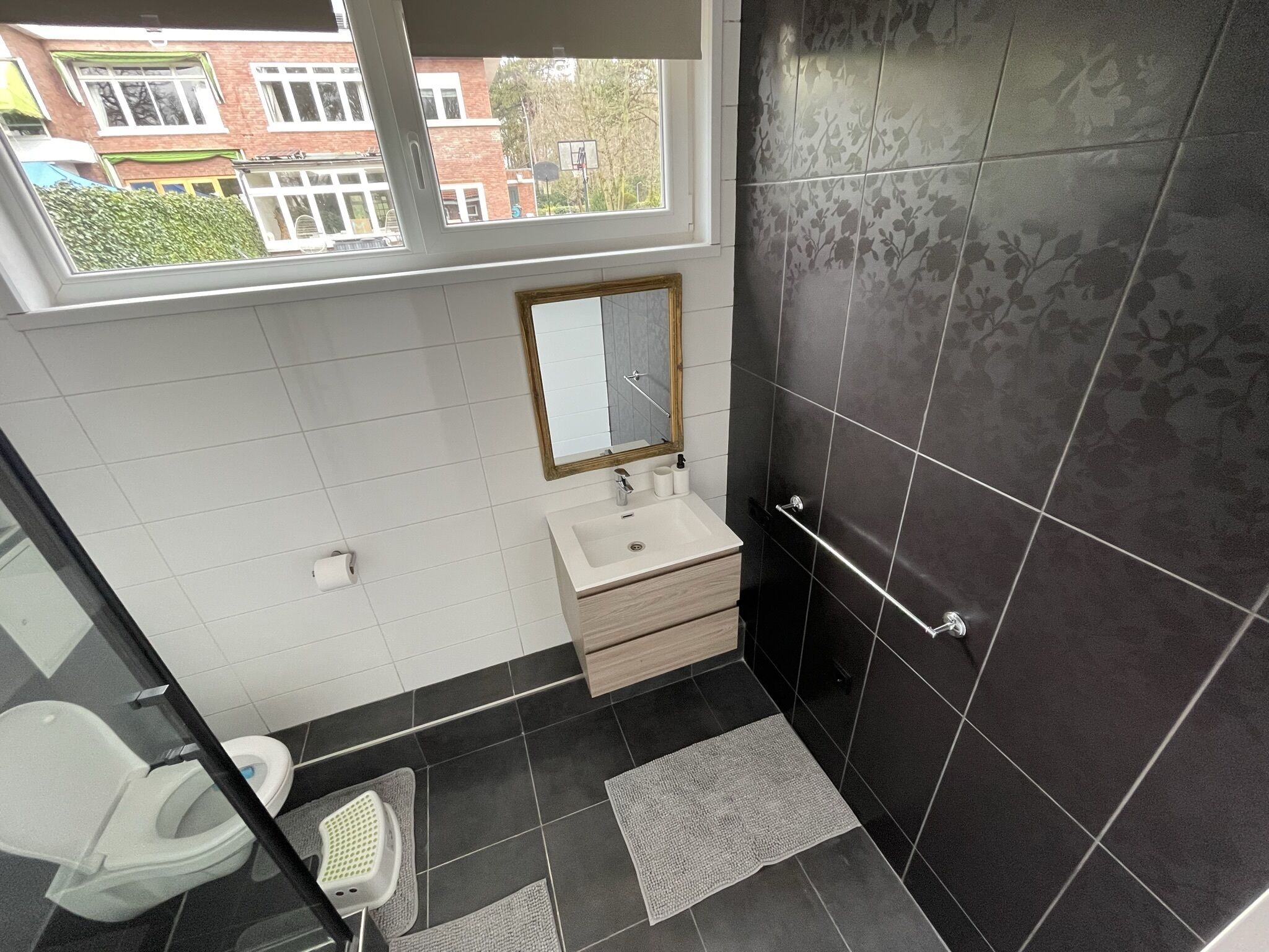 Et moderne badeværelse med toilet, håndvask og bruser. Der er et vindue, der giver naturligt lys og udsigt ud.