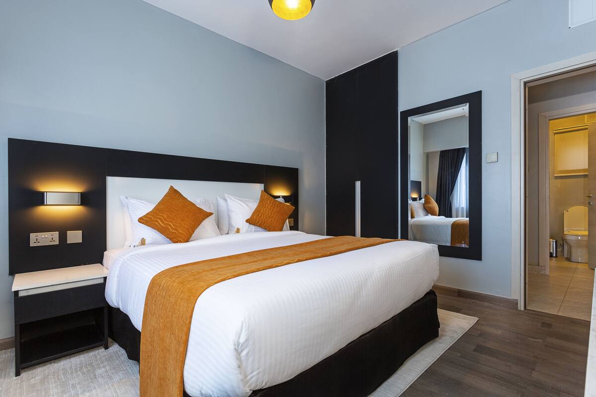 A modern hotel room with a large bed, bedside tables, a nightstand, a mirror, and a bathroom visible through an open door.