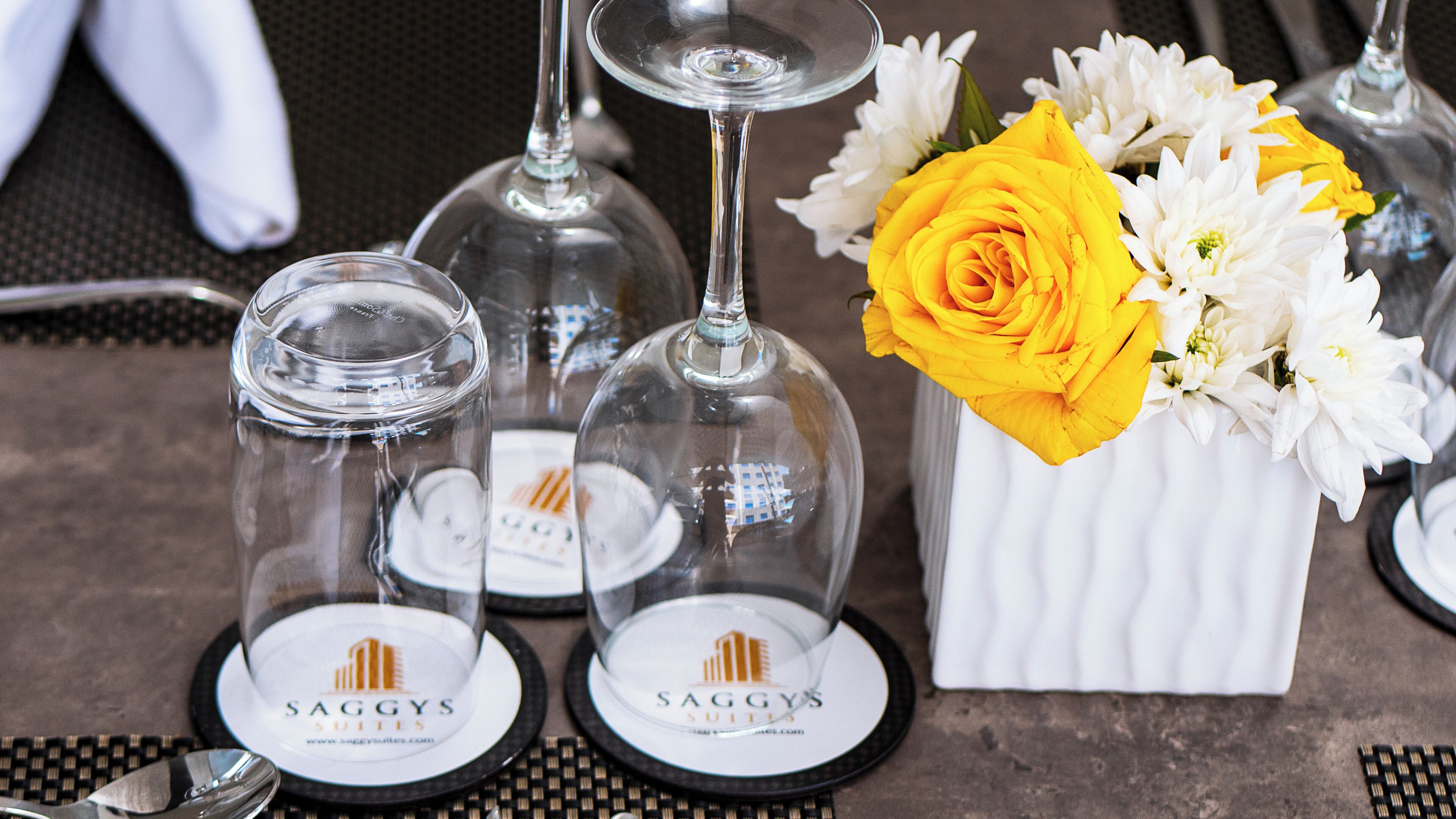 A table setting with wine glasses, a vase with a yellow rose, and a white vase with white flowers.