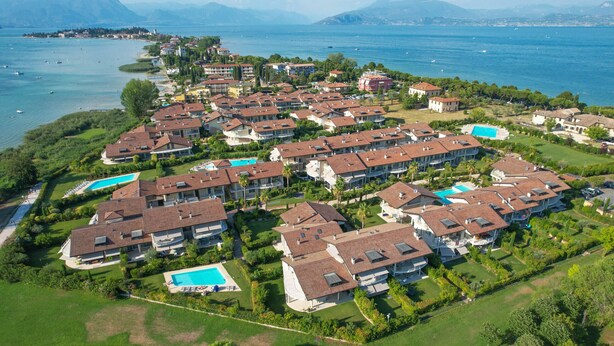 Vista aérea de una zona residencial costera con varias casas, algunas con piscinas, y un amplio cuerpo de agua y montañas al fondo.