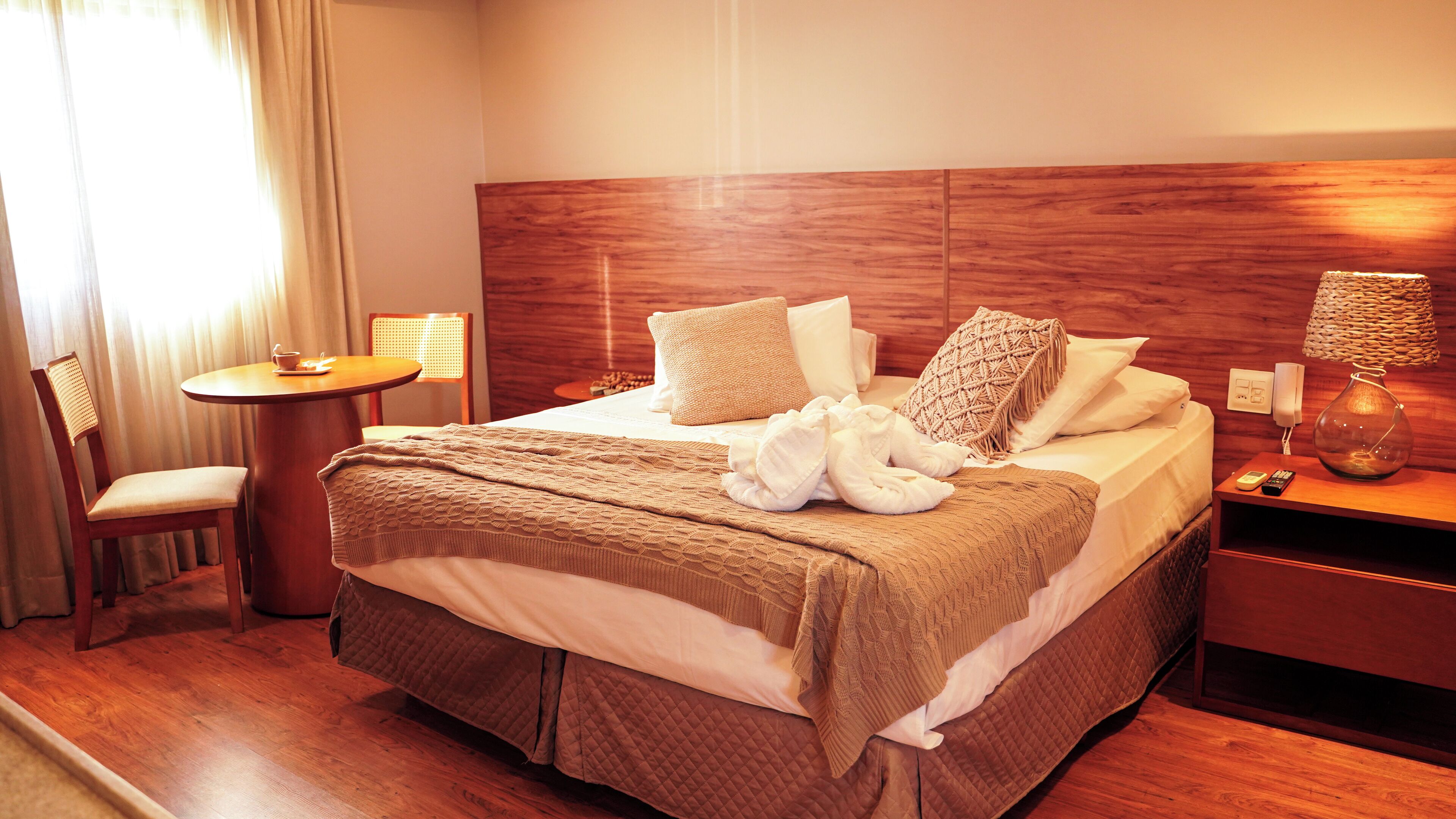 A hotel room with a wooden headboard, a round table with a chair, a bedside table with a lamp, and a bed with a quilted bedspread and pillows.