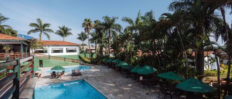 A swimming pool with lounge chairs and umbrellas, surrounded by palm trees and a building with a red roof.
