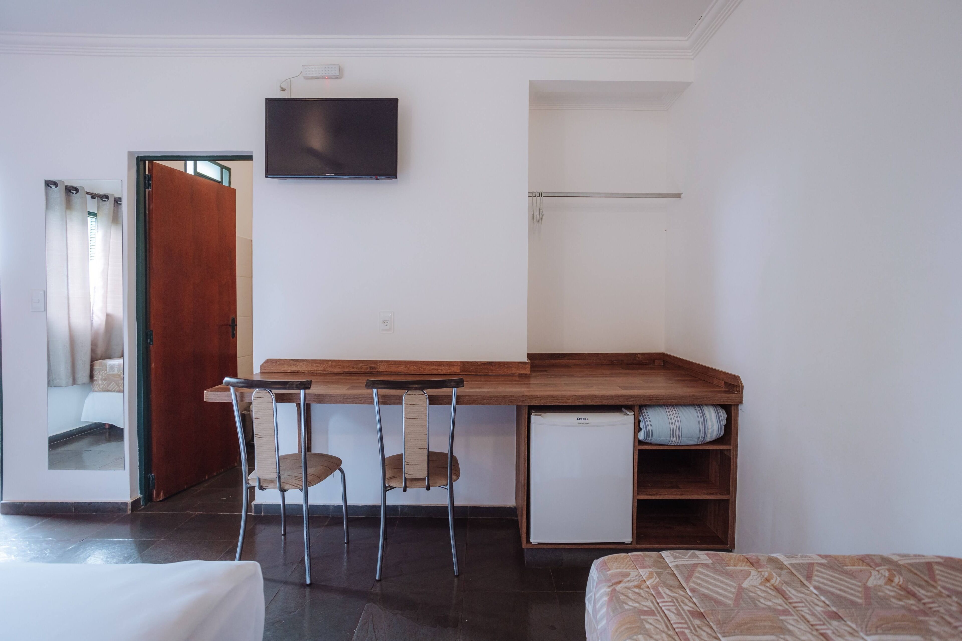 Quarto compacto com escrivaninha de madeira, duas cadeiras, um frigorífico pequeno, uma cama e uma televisão na parede.