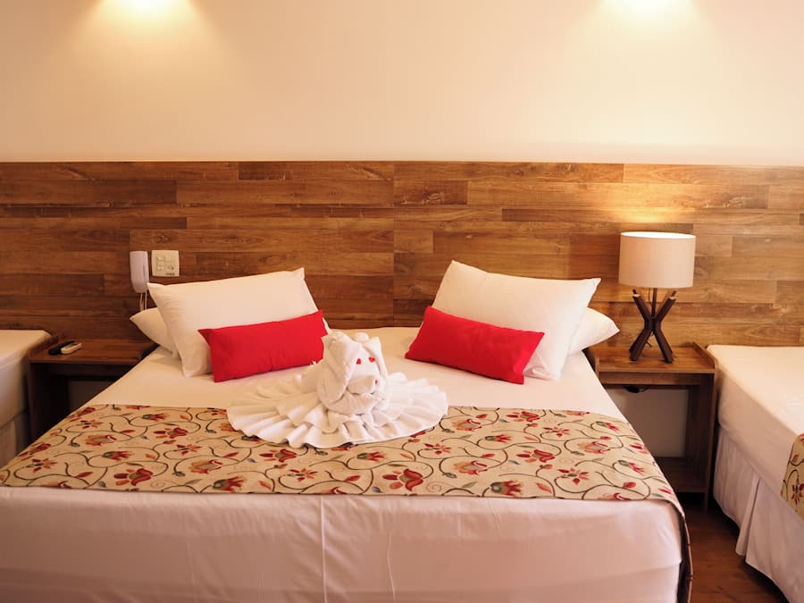 Habitación de hotel con dos camas, cada una con ropa de cama blanca y almohadas rojas. Se observa un cabecero de madera, una lámpara de noche y una colcha con estampado floral.