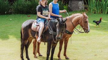 Two people riding horses in a grassy area with trees and a rooster in the background.