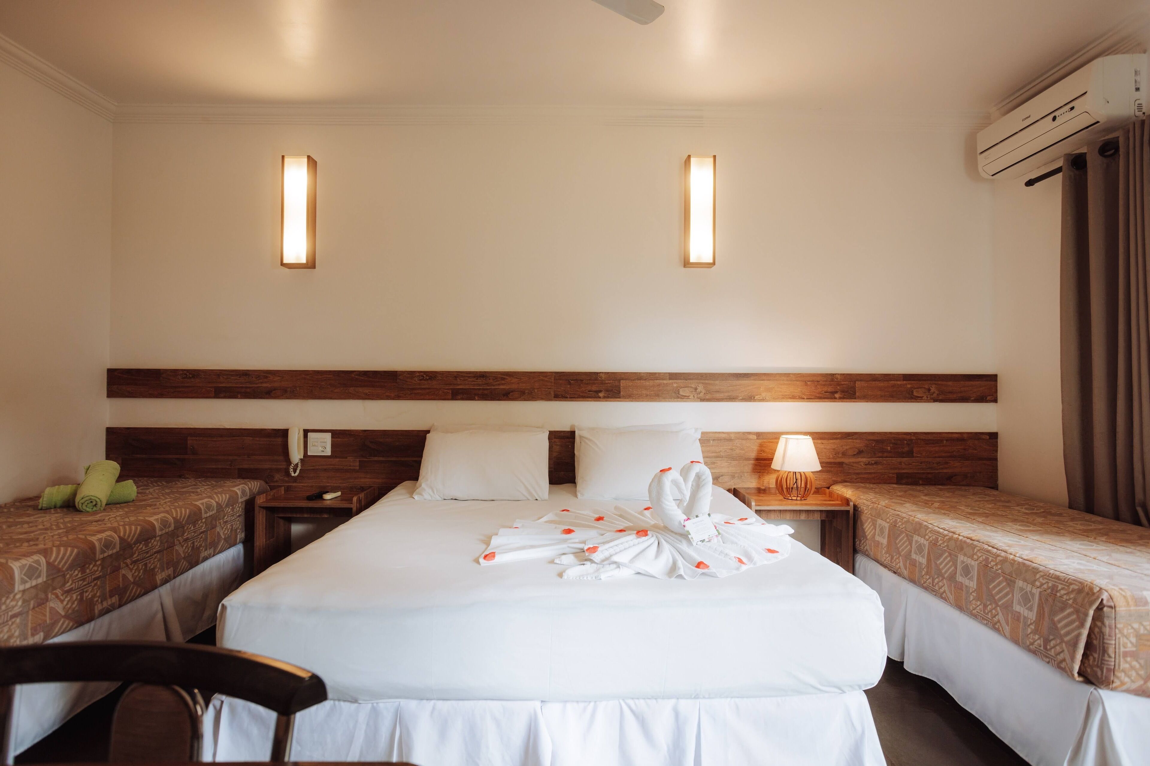 A hotel room with two single beds, a wooden headboard, a desk with a lamp, and an air conditioning unit.