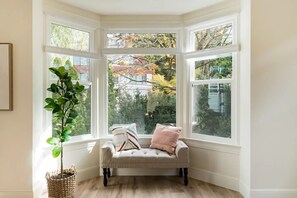 Relax in a cozy corner room with a bay window overlooking a residential area, featuring a plush bench and a large leafy plant.