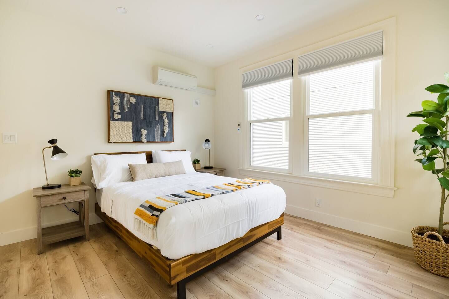 Une chambre avec un lit en bois, du linge de lit blanc, une grande fenêtre avec des stores, un climatiseur fixé au mur et une plante en pot.