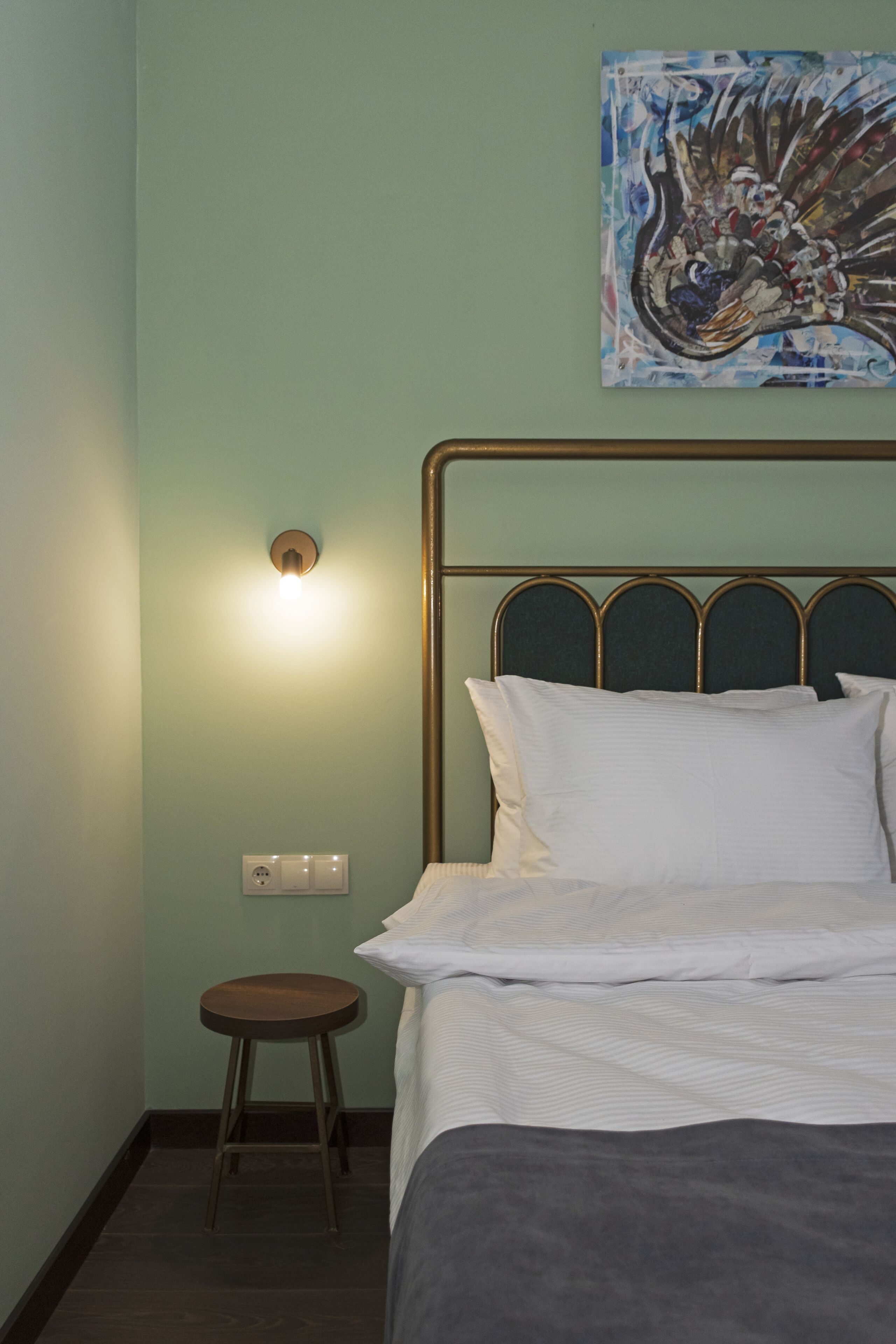 A neatly made bed with white linens and a gray blanket, a small wooden stool, and a wall-mounted light fixture.