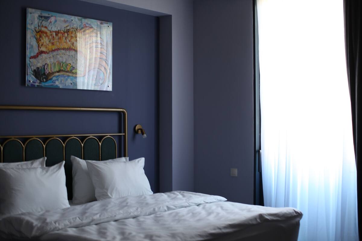 Ein Schlafzimmer mit einem Bett, einem Kopfteil, einem Bild an der Wand und einem Fenster mit Vorhängen.