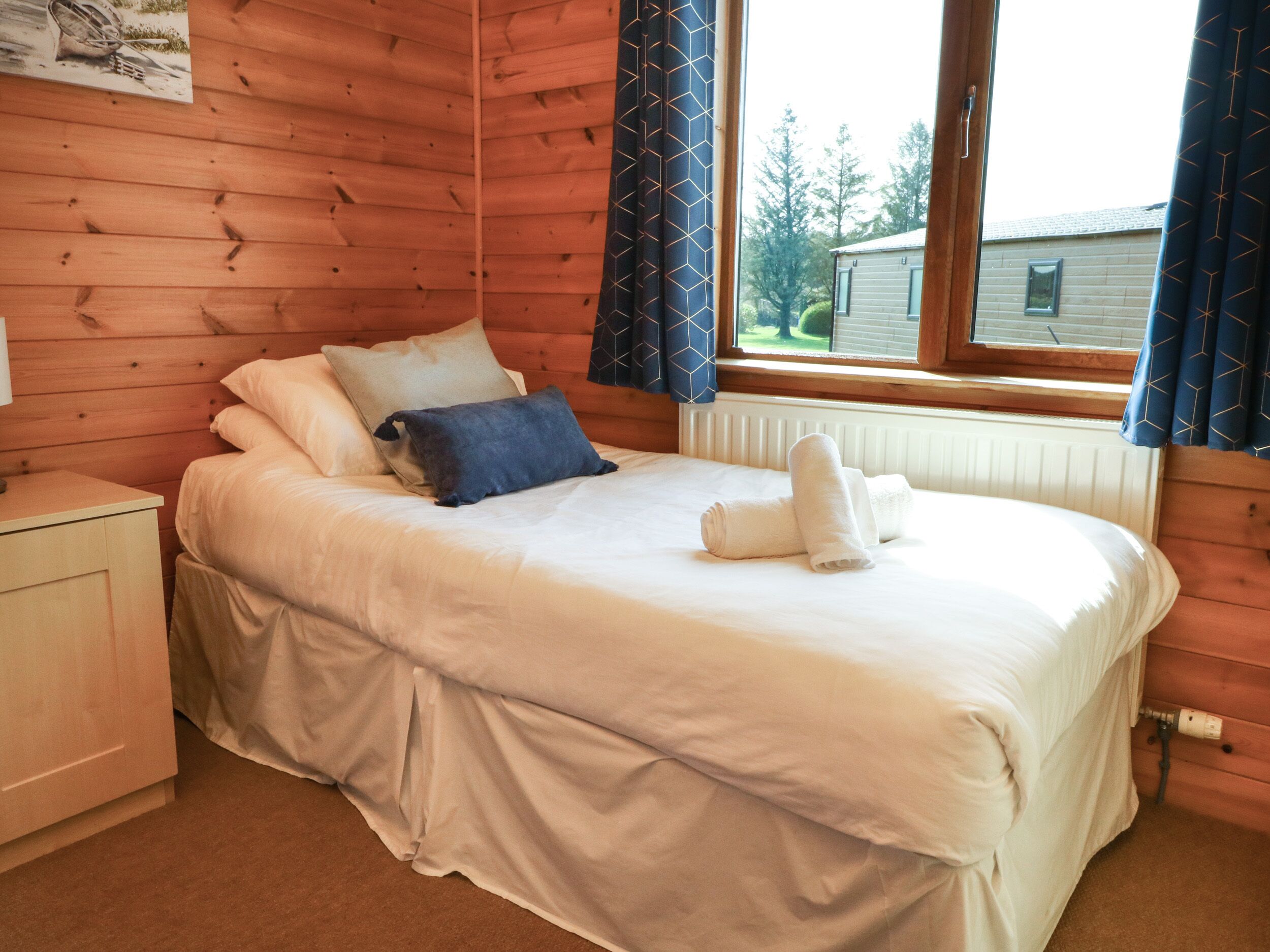 A wooden cabin bedroom with a bed, pillows, a nightstand, and a window with curtains.