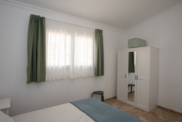Une pièce comprenant un lit, une armoire, un petit tabouret et une fenêtre avec des rideaux.